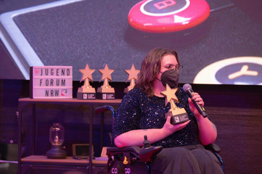 Eine Frau in einem Rollstuhl, die einen Gaming ohne Grenzen Award und ein Mikrofon hält. 